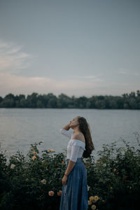 Side view of young woman standing by plants against sky