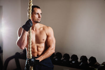 Shirtless muscular man holding rope while standing in gym