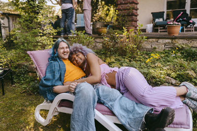 Happy gay man with transwoman lying down on chair during party in back yard