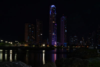 Illuminated buildings by river against sky at night