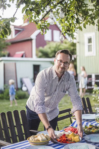 Portrait of mature man placing food plate on table in backyard while preparing for garden party