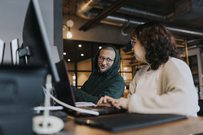 Young male hacker discussing with female colleague at desk in creative workplace