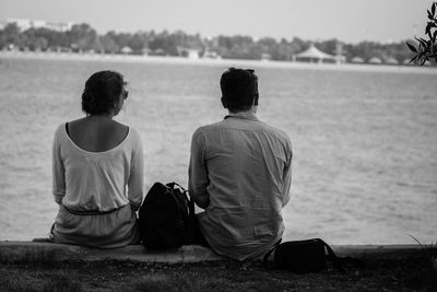 Rear view of couple sitting on retaining wall by sea