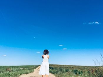 Rear view of woman standing on dirt road against blue sky