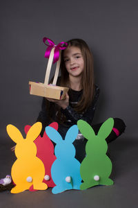 Portrait of smiling girl with eater bunny and pom pom in basket against gray background