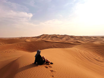Rear view of woman sitting on sand at desert against sky during sunset