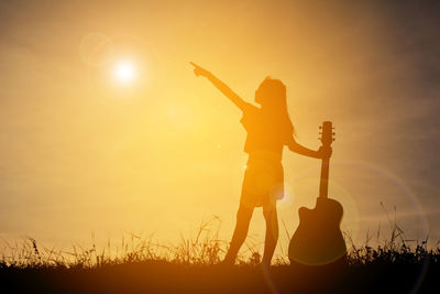 Silhouette girl with hand raised holding guitar while standing on field during sunset