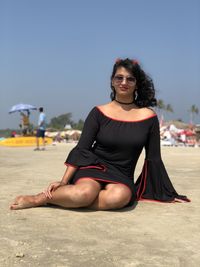 Portrait of young woman wearing sunglasses and black dress sitting at beach during sunny day