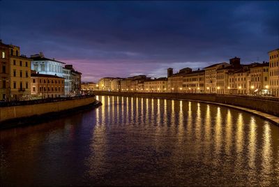 Illuminated buildings by river against sky at sunset