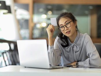 Woman holding visiting card while sitting by laptop on table in office