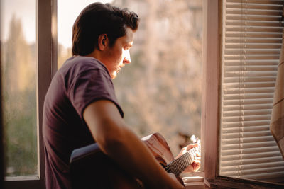 Young man playing guitar while looking out of the window