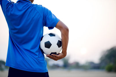 Midsection of man holding soccer ball standing on soccer field against sky