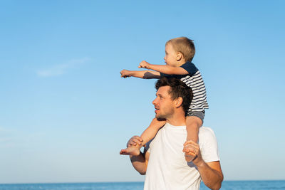 Father carrying son on shoulders at beach against clear blue sky