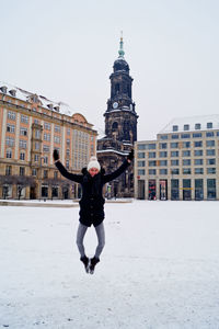 Full length of woman with arms raised jumping over snowy field against church