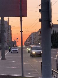 Traffic on city street by buildings against sky during sunset