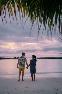 Rear view of friends standing on beach against sky during sunset