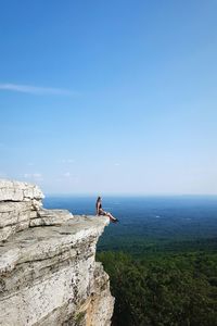 Side view of young woman sitting on cliff by landscape against blue sky