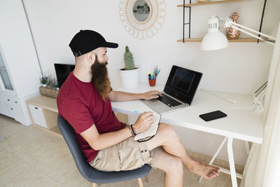 Bearded editor using laptop computer while writing on diary at home