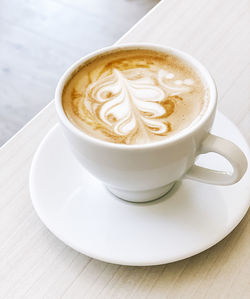 High angel view of fresh cappuccino coffee with latte art in white ceramic cup on plate on table.