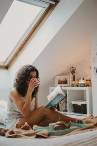 Woman drinking coffee while reading book on bed