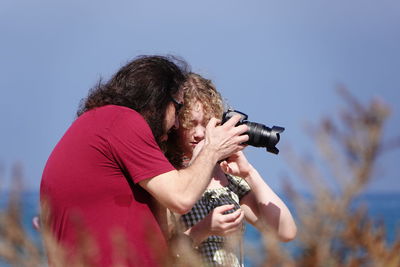 Side view of a man photographing woman against sky