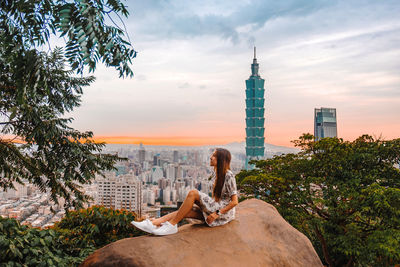 Man relaxing by buildings in city against cloudy sky