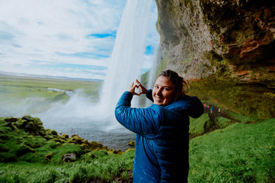 Portrait of smiling woman making heart shape against waterfall