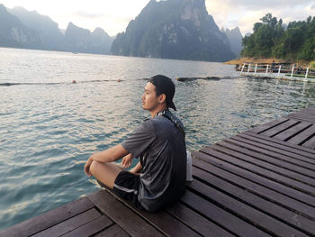 Man sitting on wooden bridge over lake against sky