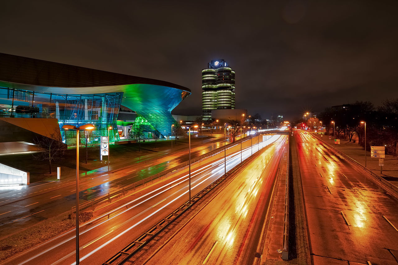 LIGHT TRAILS ON ROAD BY ILLUMINATED BUILDINGS AT NIGHT