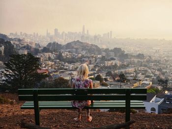 Rear view of girl looking at cityscape while sitting on bench against clear sky