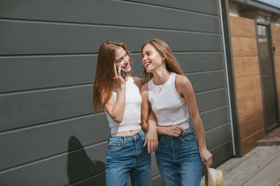 Generation z twin sisters walking outdoors, laughing while one of them talking on phone.