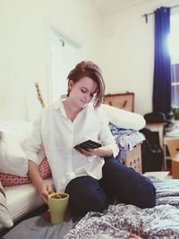 Young woman using mobile phone while sitting with tea cup at home