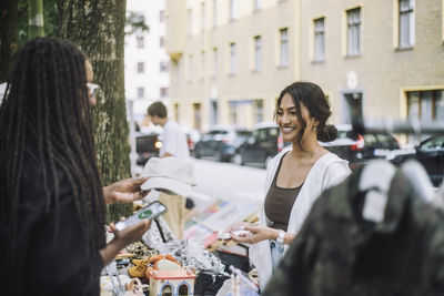 Woman buying cap from stall while talking to smiling female vendor at flea market