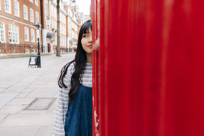 Young woman with long hair standing behind telephone booth in city