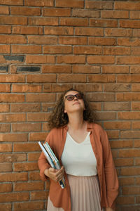 Young adult millennial with curly hair looks to sky. red brick wall. copy space. student holds books