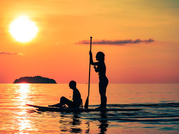 2 young women paddle boarding on sea against sunset