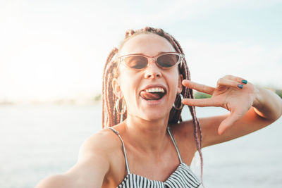 Portrait of young woman wearing sunglasses and gesturing against sky