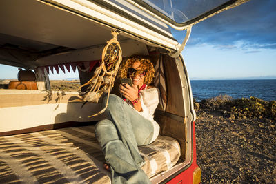 Smiling woman suing mobile phone sitting in car trunk