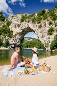 Couple sitting on picnic blanket at beach against rock formation