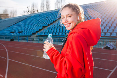 Portrait of smiling woman drinking water while listening music on running track