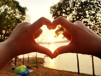 Cropped hands of person making heart shape against sky during sunset