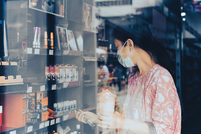 Woman in protective mask shopping in beauty and make up store shot through window.