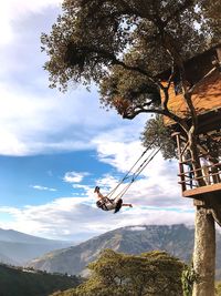 Full length of carefree woman swinging on rope swing against mountains and cloudy sky