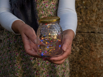 Midsection of woman holding string lights in jar against stone wall