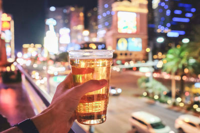 Cropped image of hand holding beer glass in illuminated city at night