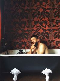 Thoughtful shirtless man sitting in bathtub against red wallpaper at home