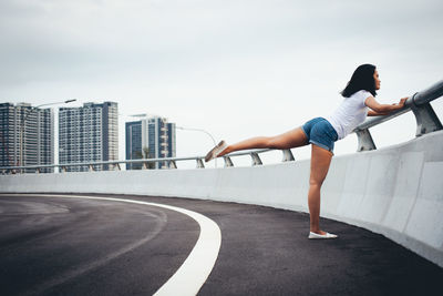 Full length side view of woman exercising by railing on bridge in city against sky