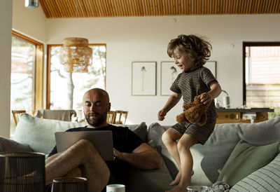 Mature male freelancer working over laptop while his son jumps on sofa and enjoys himself at home