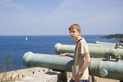 Portrait of young man standing by cannon against sea