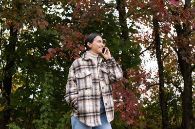 A girl in warm clothes is talking on the phone against the background of the forest and laughing